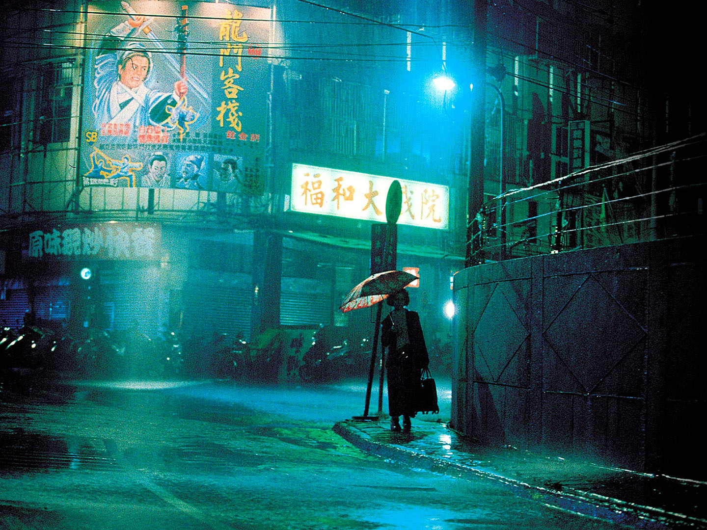 still from Goodbye, Dragon Inn (2003) - a rainy night on a taipei street lit in green and blue, a marquee advertising the screening of Dragon Inn is in the background, a woman walks with a red umbrella with white flower designs in the foreground.